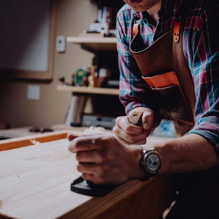 A person shaping wood with a hand plane on a workshop bench.